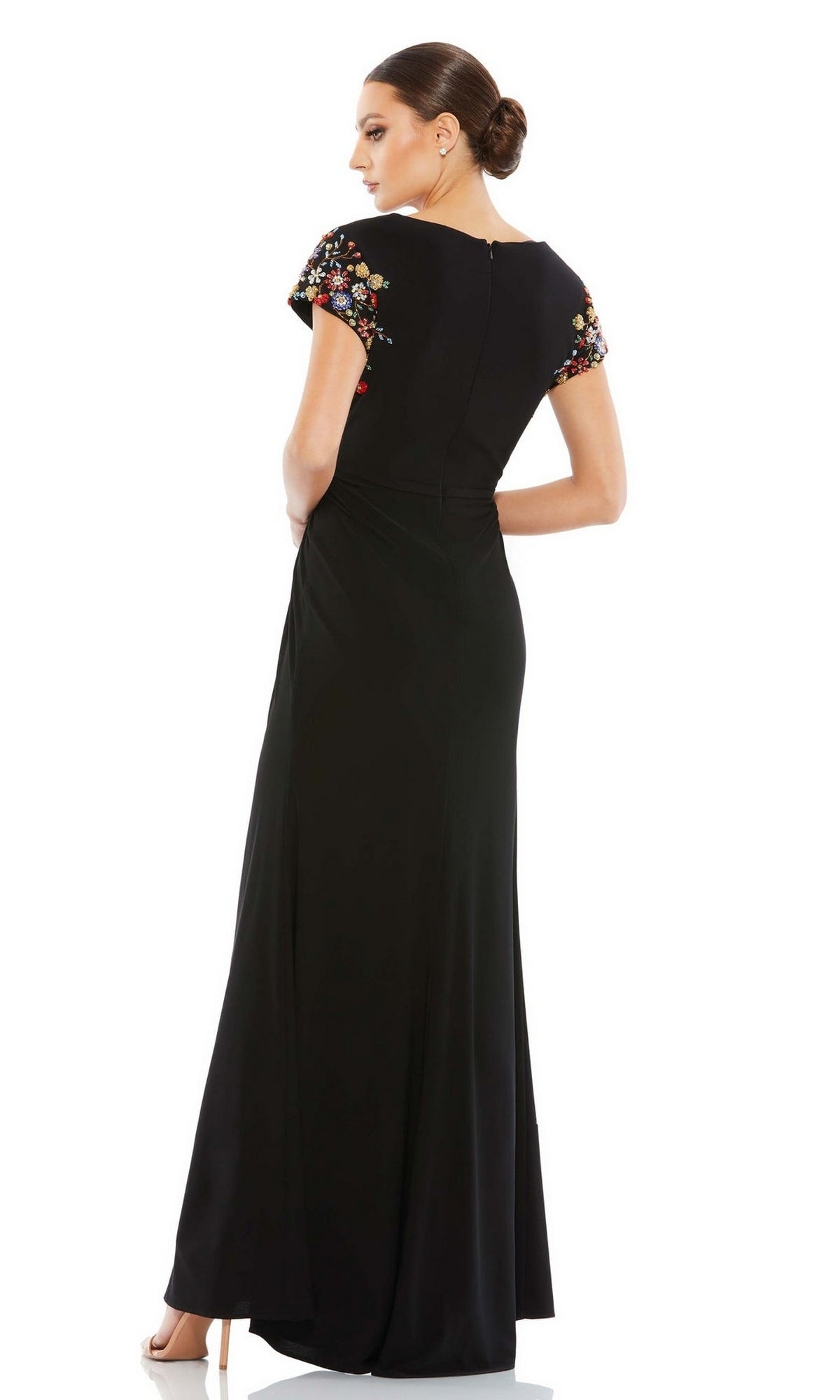 Woman in a floor-length black Cap-Sleeve Long Formal Gown (Mac Duggal 55709) with colorful floral embroidery, short sleeves, and hair styled in a low bun, standing with her back turned.